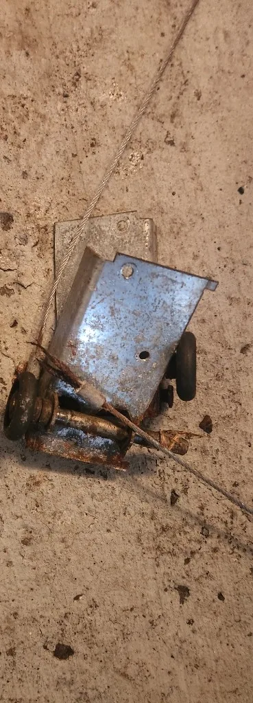 Old, heavily rusted garage door bottom bracket and roller removed from a Nepean door, with the lift cable still attached, lying on a concrete floor